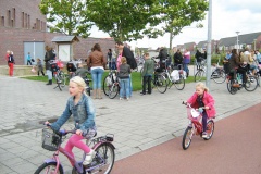 Two young girls ride home from school using a cycle path. Other children and parents stand and talk by their bikes on the footpath in the background.